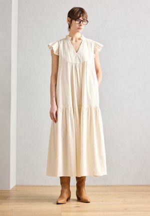 Robe fluide à volants, de couleur beige clair, avec des manches à volants en forme de cap, un décolleté en V et un tissu texturé. Associée à des bottines brunes.