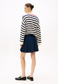 Gestreifter marineblauer und weißer Pullover mit rotem Ausschnitt, kombiniert mit einem plissierten marineblauen Rock und schwarzen Slippern, von hinten betrachtet.