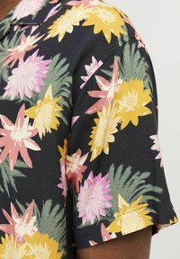 Camisa de manga corta de tela negra con un vibrante patrón floral que incluye flores amarillas, rosas y blancas, complementadas con hojas verdes.