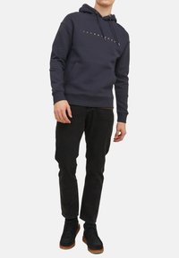 Marine hoodie met koord en voorzak, met gouden logo tekst. Gekocht bij zwarte jeans en donkere schoenen met bruine zolen.
