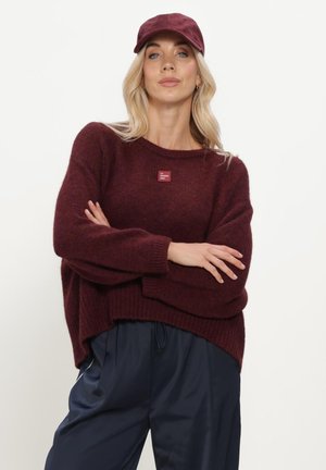 Frau mit blondem Haar trägt eine burgunderfarbene Kappe, passenden Strickpullover mit Etikett und lockere dunkelblaue Hose und steht mit verschränkten Armen.