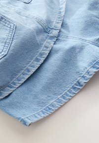 Chapeau bob en denim bleu clair avec des coutures visibles et une partie d'une poche avant sur un fond clair.
