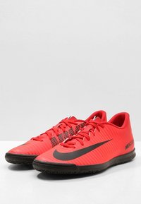 Zapatillas deportivas rojas hechas de material sintético, con un diseño acanalado, detalles en negro y una suela negra texturizada con un logo destacado.