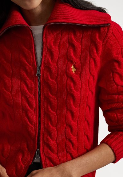 Czerwony sweter w splot kablowy z wysokim kołnierzem i zamkiem z przodu. Małe złote logo na piersi, teksturowany wzór oraz ściągacze na mankietach.