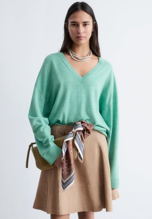 Kvinde iført mintgrøn V-hals sweater, tan nederdel med mønstret tørklæde bundet om livet, holder tan håndtaske og lagdelte perlekæder.