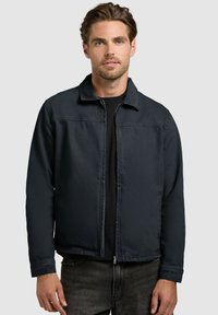 Młody mężczyzna w czarnej kurtce na zamek błyskawiczny nałożonej na czarną koszulę, stojący na tle jednolitego jasnoszarego tła.