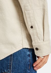 Beige cotton shirt with a structured collar, button closure at the cuff, and visible stitching; paired with blue denim jeans.