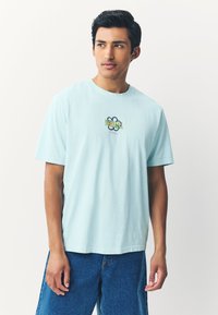 Hellblaues Baumwoll-T-Shirt mit Rundhalsausschnitt und kurzen Ärmeln, versehen mit einem mehrfarbigen grafischen Design vorne. Jeans darunter sichtbar.