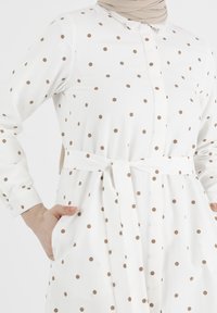 Robe blanche à pois beiges, manches longues, avec un devant à boutons, une ceinture nouée à la taille et des poches latérales. Texture de tissu lisse.
