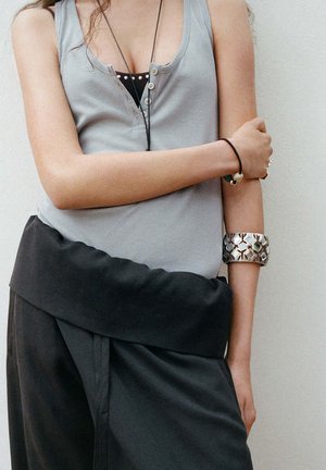 Person mit verschränkten Armen, die ein graues ärmelloses Oberteil, schwarze Hose, ein silbernes Manschettenarmband und ein schwarzes Haargummi am Handgelenk trägt.