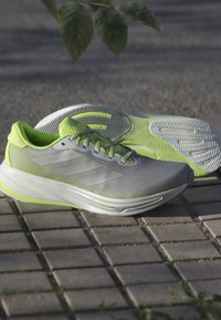 Buty sportowe z szarym siateczkowym wierzchem, zielonymi akcentami i teksturowaną białą podeszwą. Elementy to sznurówki oraz trzy białe paski po boku.