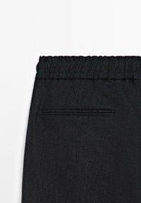 Zwarte stoffen broek met elastische tailleband en een enkele paspelzak op de achterkant, weergegeven tegen een witte achtergrond.