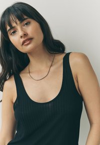 Sort ribstrikket tanktop med rund halsudskæring og tynde stropper, båret med en guldfarvet snoet kædehalskæde. Glat tekstur, tætsiddende design.