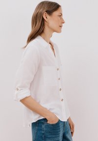 Camicia bianca con bottoni, realizzata in tessuto leggero, caratterizzata da una vestibilità rilassata, maniche lunghe, una tasca sul petto e bottoni color oro.