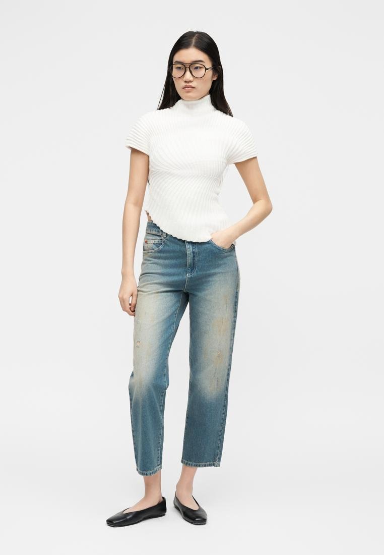 Femme portant des lunettes noires, un haut à col roulé côtelé blanc à manches courtes, un jean bleu délavé et des ballerines noires, debout avec une main dans la poche.
