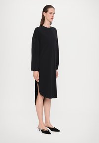 Kvinna iklädd en långärmad svart midikjol med slit på sidan, i kombination med svarta spetsiga slingback-klackar, stående mot en enfärgad ljus bakgrund.