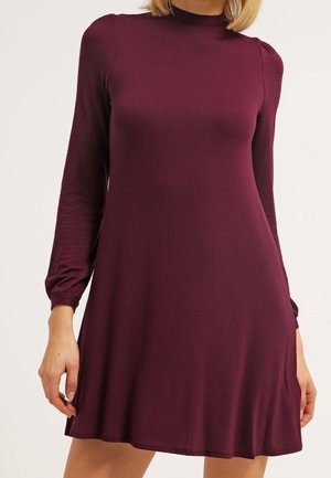 Kobieta w bordowej, długorękawowej sukience w kształcie litery A z wysokim kołnierzem, marszczonymi ramionami i mankietami na jednolitym tle.
