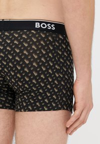Μαύρα ανδρικά εσώρουχα μποξεράκια με γεωμετρικό σχέδιο σε μπεζ, με μαύρη ελαστική ζώνη μέσης που φέρει το λογότυπο "BOSS" σε λευκό.