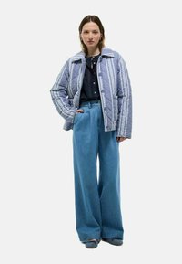 Veste matelassée bleu clair avec des rayures blanches verticales, dotée d'un large col et de boutons sur le devant. Associée à un pantalon en denim bleu ample.
