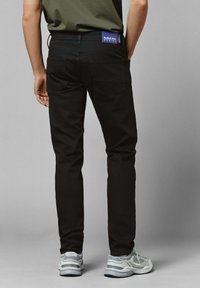 Henkilö, joka käyttää mustia slim fit -farkkuja, joissa on Red Bull Racing -logo vyötäröllä, ja valkoisia lenkkikenkiä, seisoo yksinkertaista taustaa vasten.