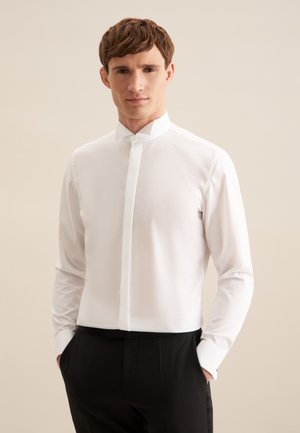 Jeune homme aux cheveux bruns courts, portant une chemise blanche à col mandarin et un pantalon noir, debout les mains dans les poches.