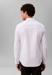 Valge pika varrukaga särk klassikalise kraega. Sile kangas kerge läikega, ümar äärejoon ja nööbitavad varrukad. Vaadeldav tagantpoolt.