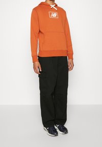 Felpa arancione con logo bianco, tasca a marsupio e cappuccio con cordino; abbinata a pantaloni cargo neri e sneakers blu navy.