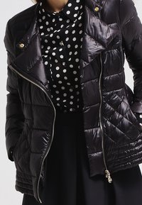 Sort quiltet dunjakke med lynlåslukning, med guldfarvede detaljer og diamantmønster, båret over en prikket bluse.