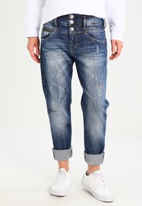 Person som har på sig uppvikt, slitna blå denimjeans med tre knappar och vita sneakers, står mot en vit bakgrund.