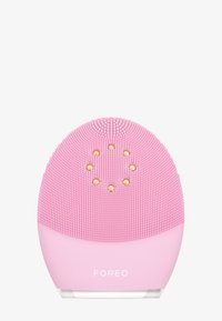 Appareil de nettoyage facial Foreo en silicone rose. Pr�ésente des nodules texturés sur le dessus et un design incurvé avec une base transparente. Accents dorés présents.