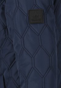 Marinblå quiltad jacka med hexagonal mönster, tillverkad av isolerat tyg, med en svart fyrkantig logotyp-patch på ärmen.