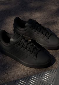 Par de zapatillas negras Adidas con cordones y las distintivas tres franjas, colocadas sobre un suelo de concreto texturizado con una rejilla metálica cercana.