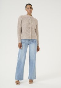 Beige getextureerde trui met knoopsluiting, gecombineerd met lichtblauwe jeans met wijde pijpen. Model draagt open teen beige hakken en staat tegen een witte achtergrond.