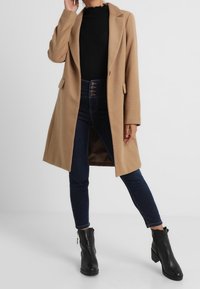 Lysebrun frakke med skræddersyet design, båret over en sort ribbet top og mørkeblå skinny jeans, parret med sorte ankelstøvler.