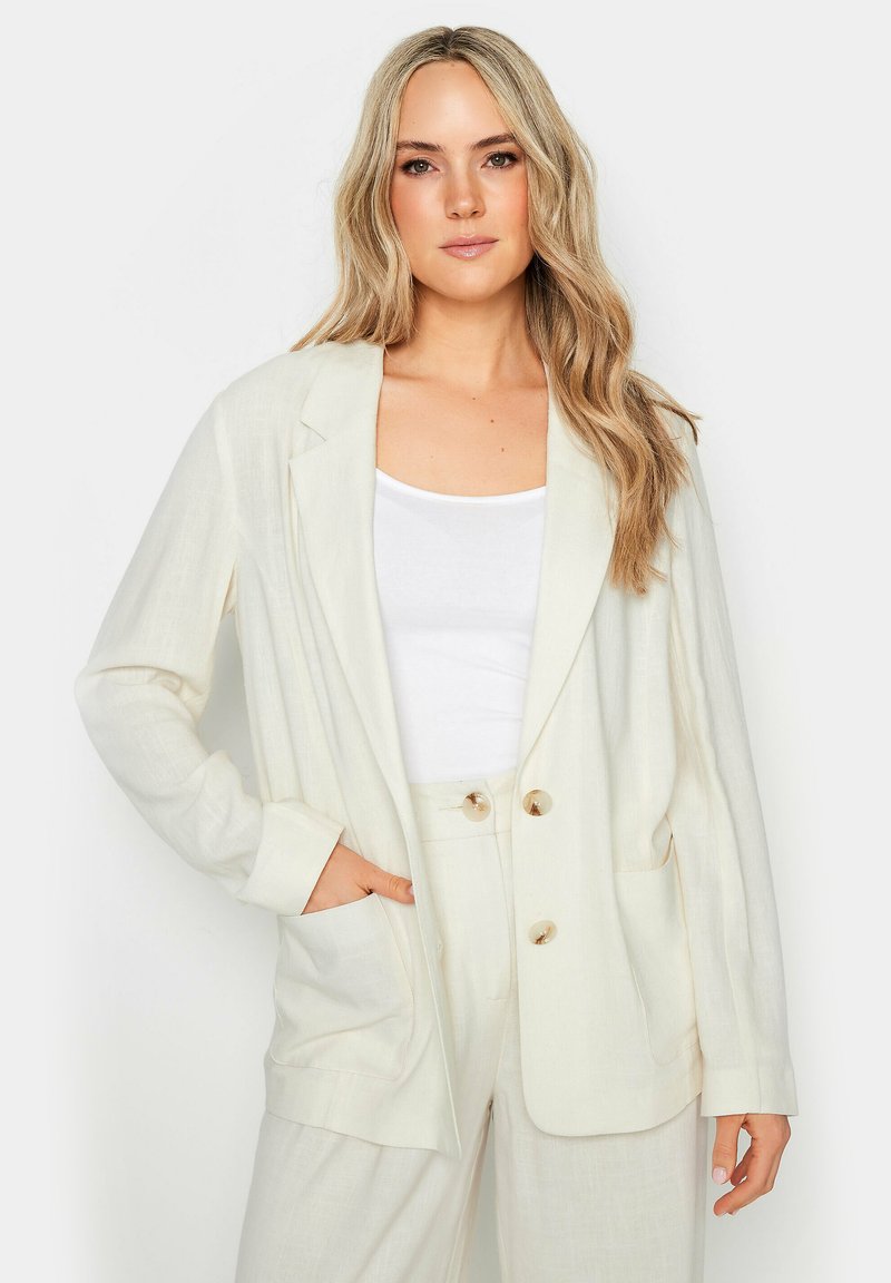 Blazer di lino color beige chiaro con due grandi tasche frontali, rever a punta e bottoni color oro. Indossato sopra una maglietta bianca e pantaloni abbinati.