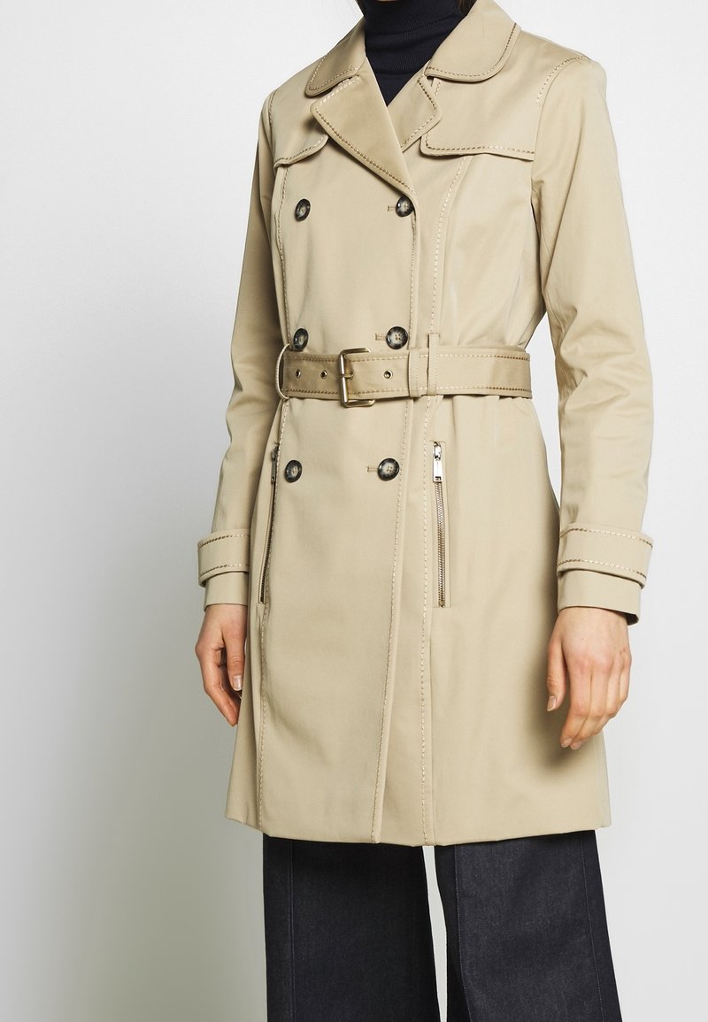 Gabardina beige con botones de doble botonadura, cintura ceñida con cinturón y bolsillos laterales con cremallera. Presenta costuras en contraste y un cuello con solapa.