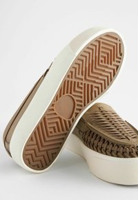 Brune vævede sneakers med en tyk hvid og tan gummisål, der har et fiskebensmønster på ydersålen og teksturerede detaljer.