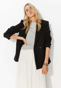 Blazer noir ajusté avec un t-shirt gris en dessous et une jupe en dentelle blanche. Le blazer a des manches roulées et deux boutons. Matériaux texturés.
