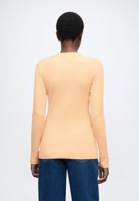 Žebrovaný top s dlhým rukávom svetlo broskyňovej farby, s okrúhlym výstrihom a priliehavým strihom. Kombinovaný s modrými džínsami s širokými nohavicami.