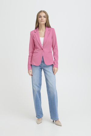 Femme portant un blazer rose, un top blanc, un jean bleu clair et des talons beiges, debout devant un fond blanc uni.