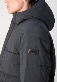 Black puffer jacket with a hood, featuring quilted texture, a smooth finish, and a logo patch on the left sleeve.