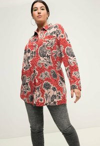 Camicia rossa a fantasia con disegni floreali neri e motivi leopardati, maniche lunghe, chiusura con bottoni, abbinata a jeans grigi. Tessuto leggero.