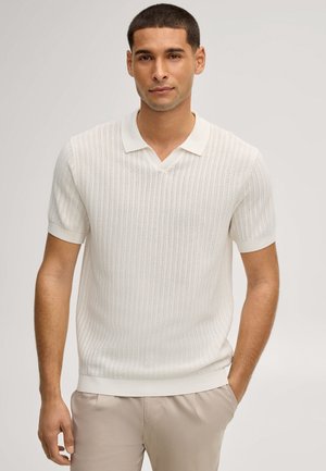 Mann trägt ein weißes, kurzärmeliges Strick-Poloshirt und beige Hose, steht mit einer Hand in der Tasche vor einfarbigem Hintergrund.