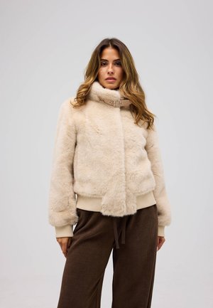 Kvinna med långt hår som bär en beige fuskpälsjacka med spännhals och bruna byxor, stående mot en enfärgad ljus bakgrund.