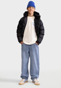 Czarna kurtka puchowa, kremowy t-shirt z niebieskim logo, jasnoniebieskie luźne dżinsy i beżowe buty z niebieskimi paskami; niebieski beanie na głowie.