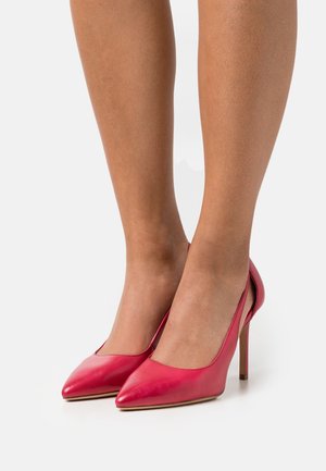 Jambes portant des escarpins aiguilles rouges brillants avec un décolleté sur fond blanc.