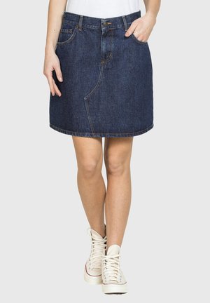 Donna che indossa una gonna di jeans blu scuro con tasche frontali e chiusura con bottoni, abbinata a sneakers bianche alte e una canotta bianca.