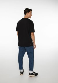 Zwarte katoenen T-shirt met een relaxed fit, korte mouwen en een ronde halslijn, gecombineerd met blauwe spijkerbroek en zwarte sneakers met witte accenten.