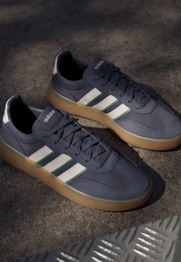 Par de zapatillas Adidas azul oscuro con rayas blancas y verdes y suelas de color marrón claro colocadas sobre una superficie de hormigón texturizado.