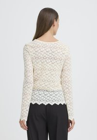 Blonde langærmet top i off-white med blomster mønster og bølget kant. Bagfra viser den let, gennemsigtig tekstur og tætsiddende design.